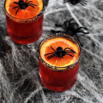 Red drink in short glasses topped with orange slice and plastic spider sitting on a spider web surrounded by small plastic spiders.