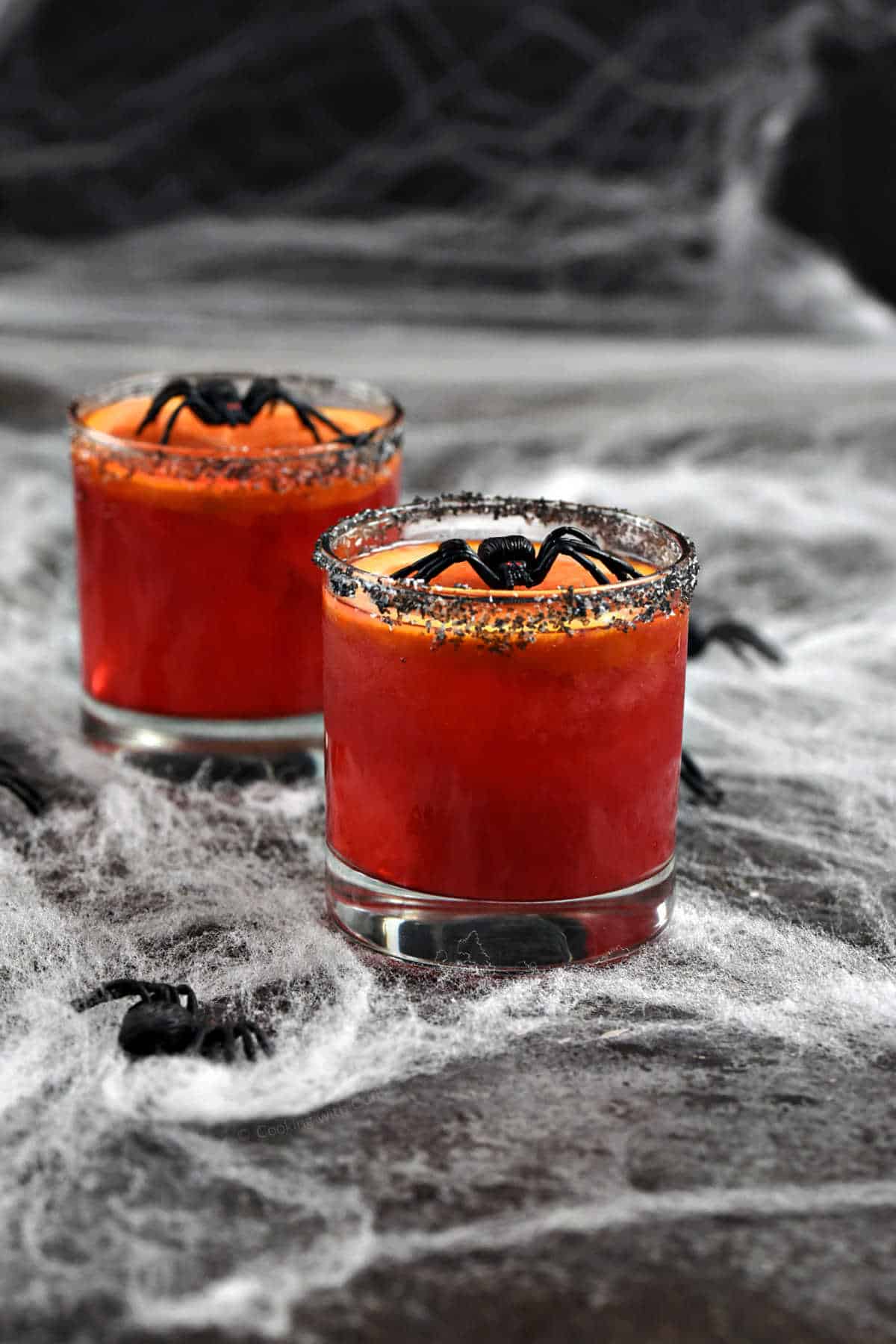 Red drink in short glasses topped with orange slice and plastic spider sitting on a spider web surrounded by small plastic spiders.