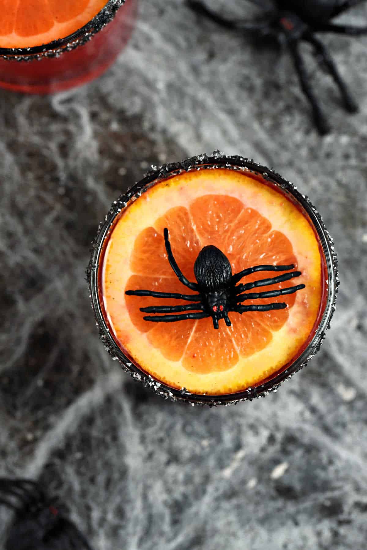 Red drink in short glasses topped with orange slice and plastic spider sitting on a spider web surrounded by small plastic spiders.