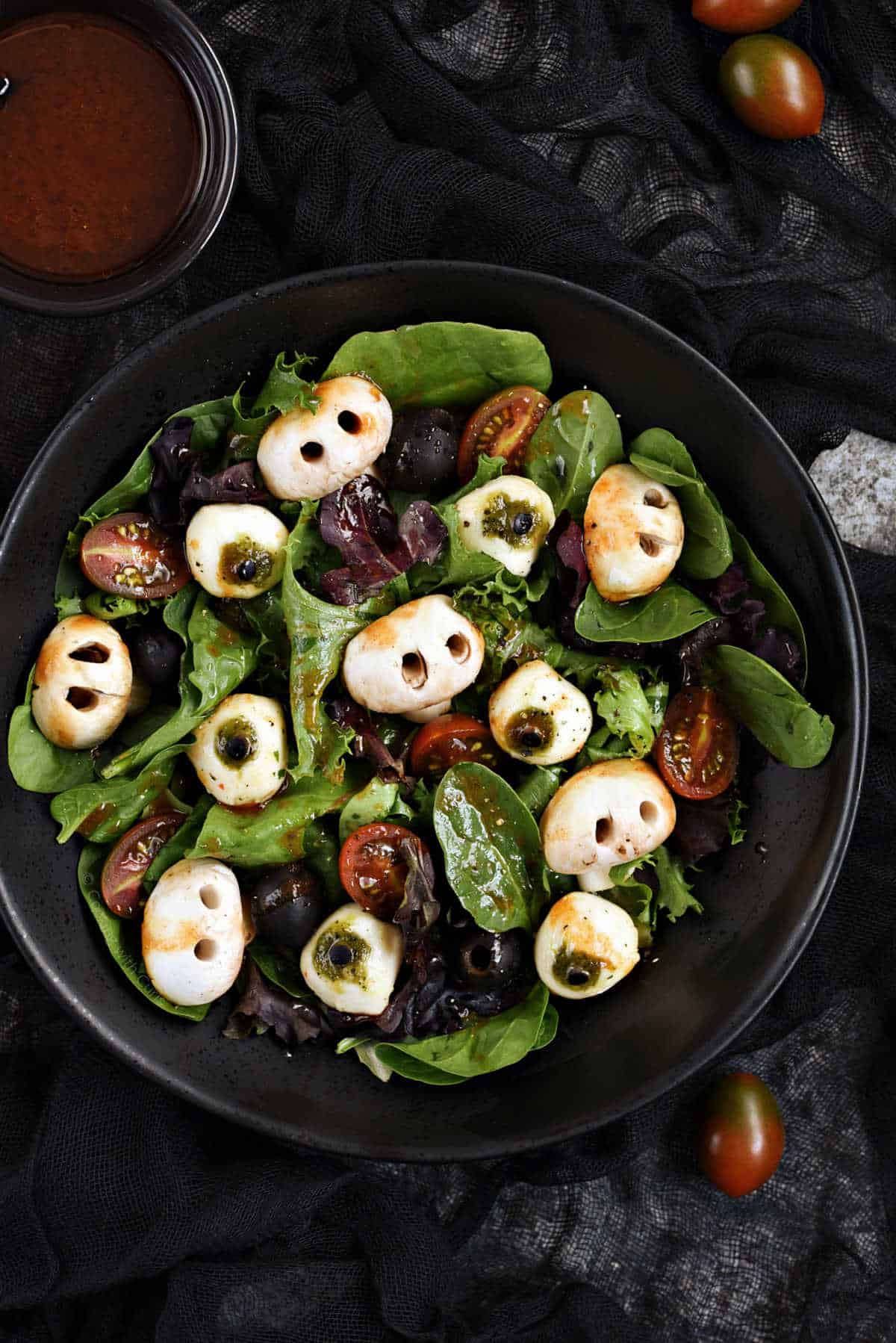 Mixed greens, mushroom skulls, mozzarella eyeballs, black olives, cherry tomatoes, and blood red vinaigrette in a black bowl.