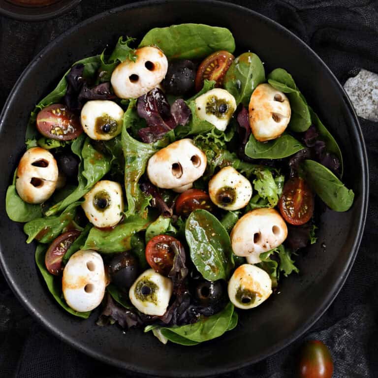 Mixed greens, mushroom skulls, mozzarella eyeballs, black olives, cherry tomatoes, and blood red vinaigrette in a black bowl.