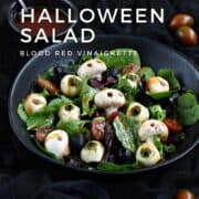 Mixed greens, mushroom skulls, mozzarella eyeballs, black olives, cherry tomatoes, and blood red vinaigrette in a black bowl with title graphic across the top.