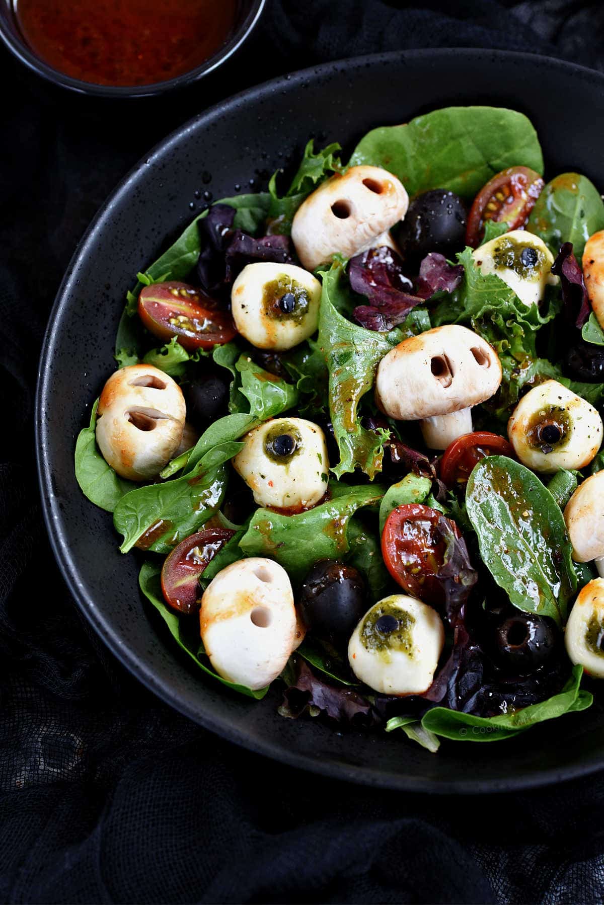 Mixed greens, mushroom skulls, mozzarella eyeballs, black olives, and blood red vinaigrette in a black bowl with red vinaigrette in a small bowl in the background.