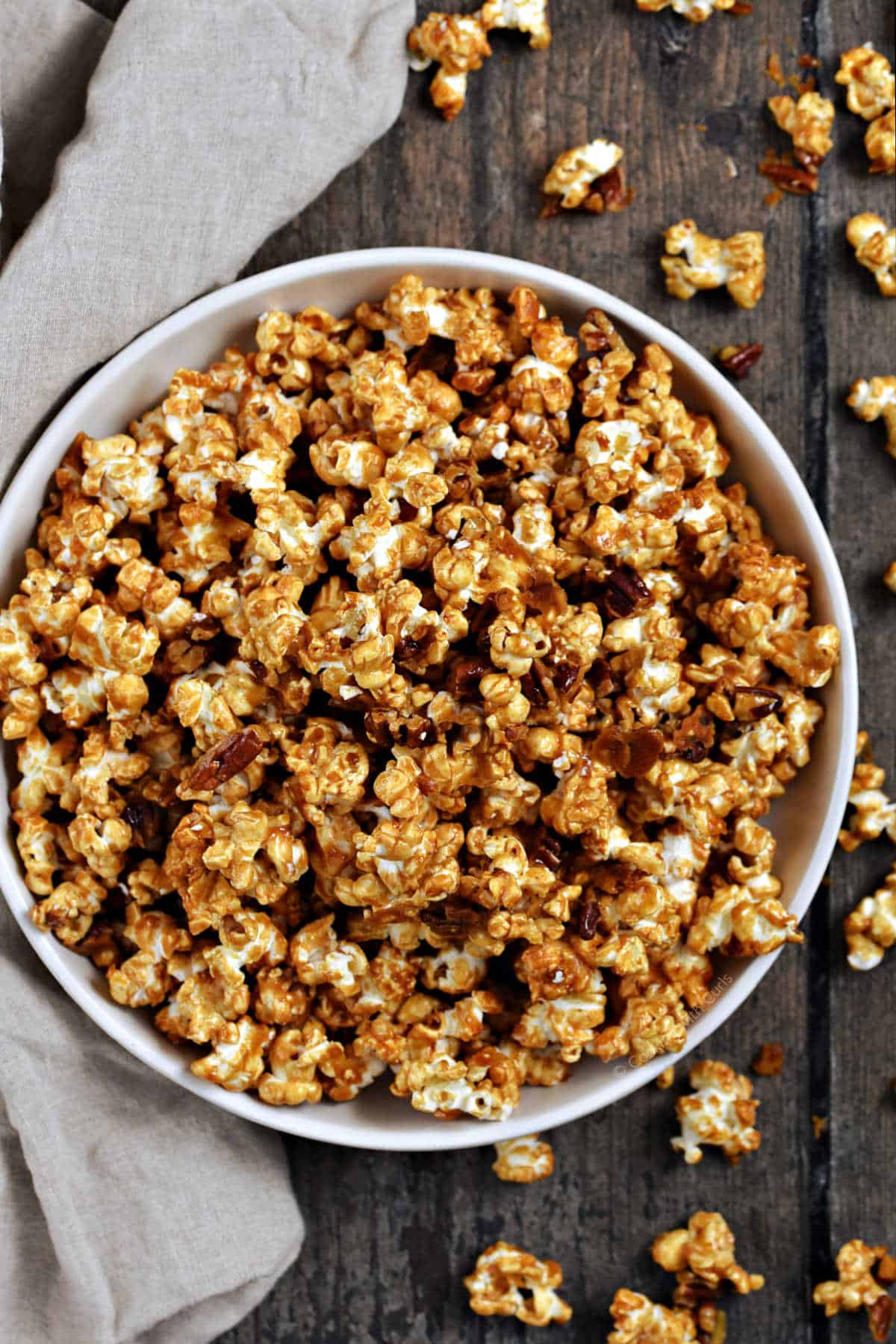 A bowl of caramel popcorn with candied pecans surrounded by scattered popcorn.