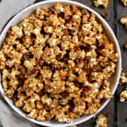 A bowl of caramel popcorn with candied pecans surrounded by scattered popcorn.