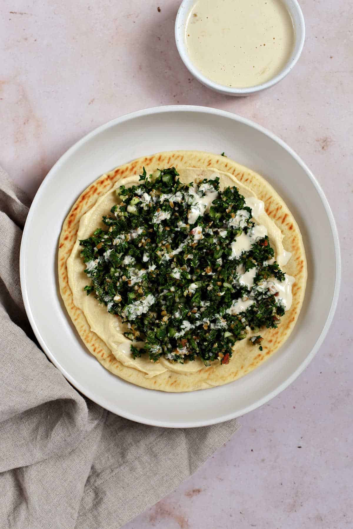 A fluffy pita bread topped with hummus, tabbouleh, and tahini sauce on a plate.