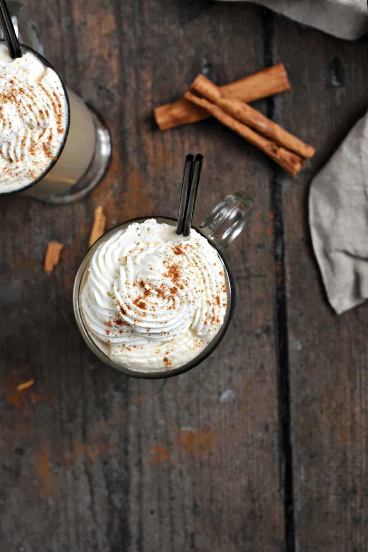 Whipped cream topped cinnamon latte cocktail in a glass mug with cinnamon and two black straws.