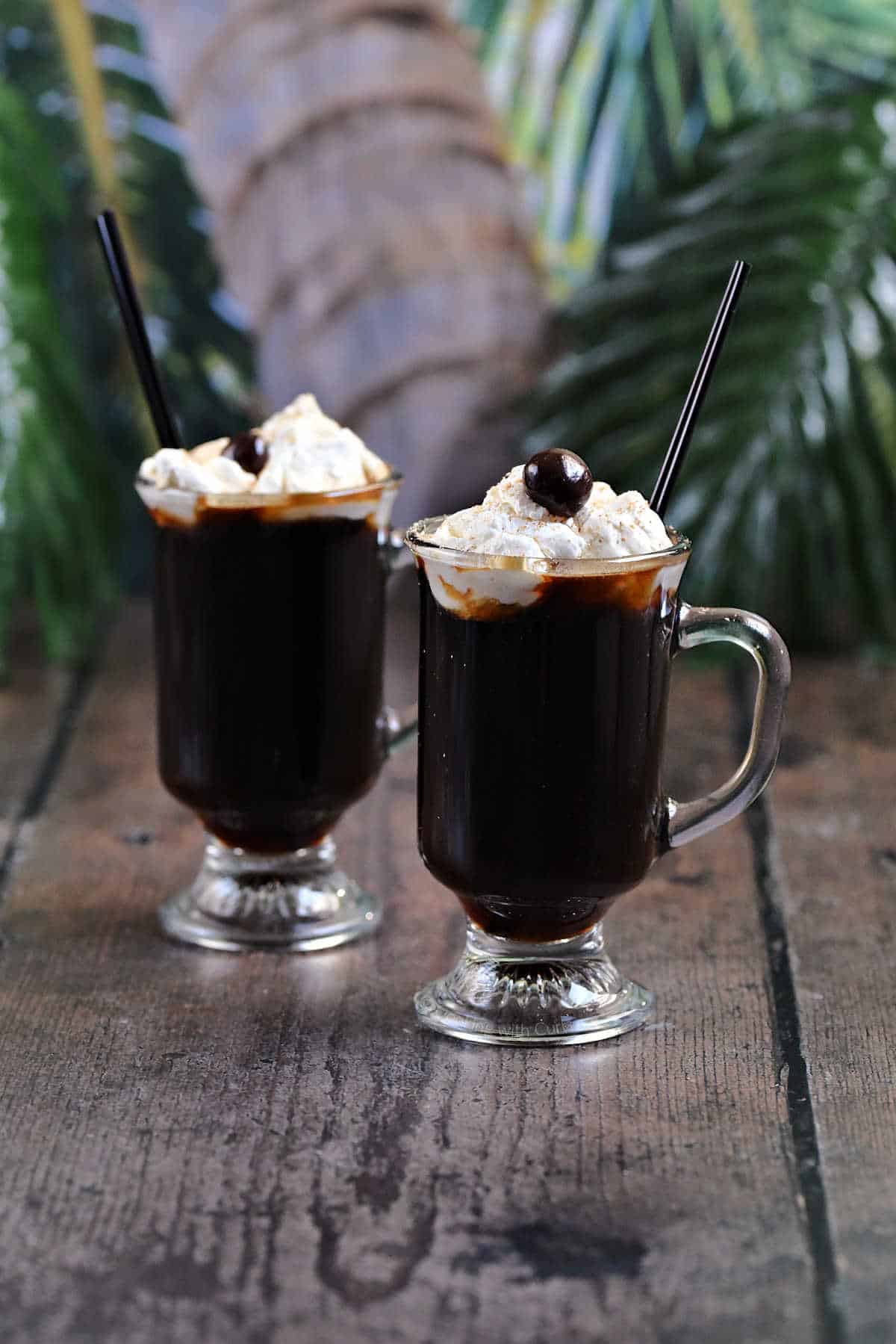 Two glass mugs filled with a coffee cocktail topped with whipped cream and a chocolate covered coffee bean with tropical leaves in the background.