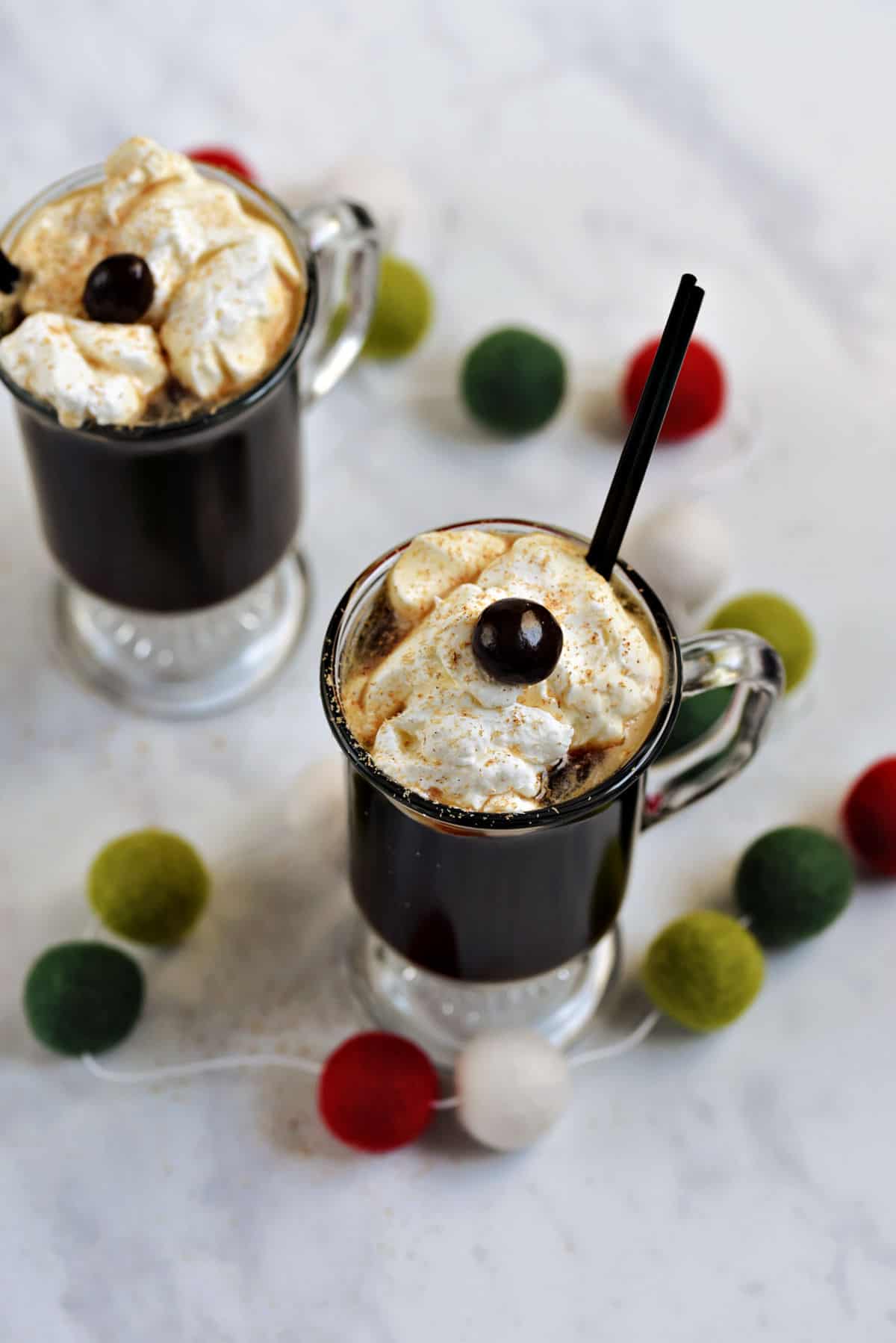 Two glass mugs filled with a coffee cocktail topped with whipped cream and a chocolate covered coffee bean surrounded by green and red wool balls.