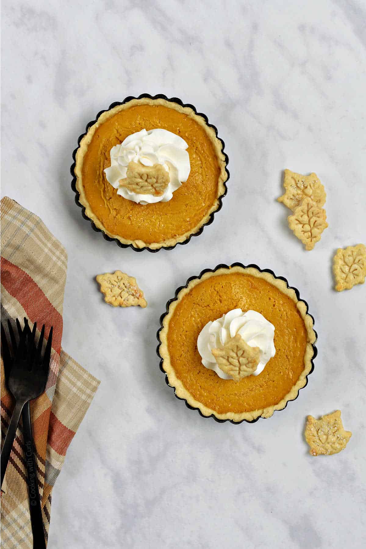 Two mini pumpkin pies with whipped cream and pie crust leaf shaped cutouts.