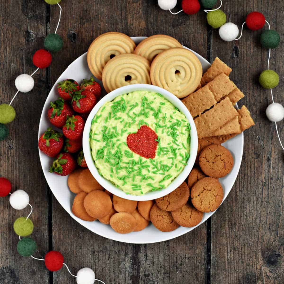 A bowl of Grinch dip on a plate surrounded by cookies and strawberries.