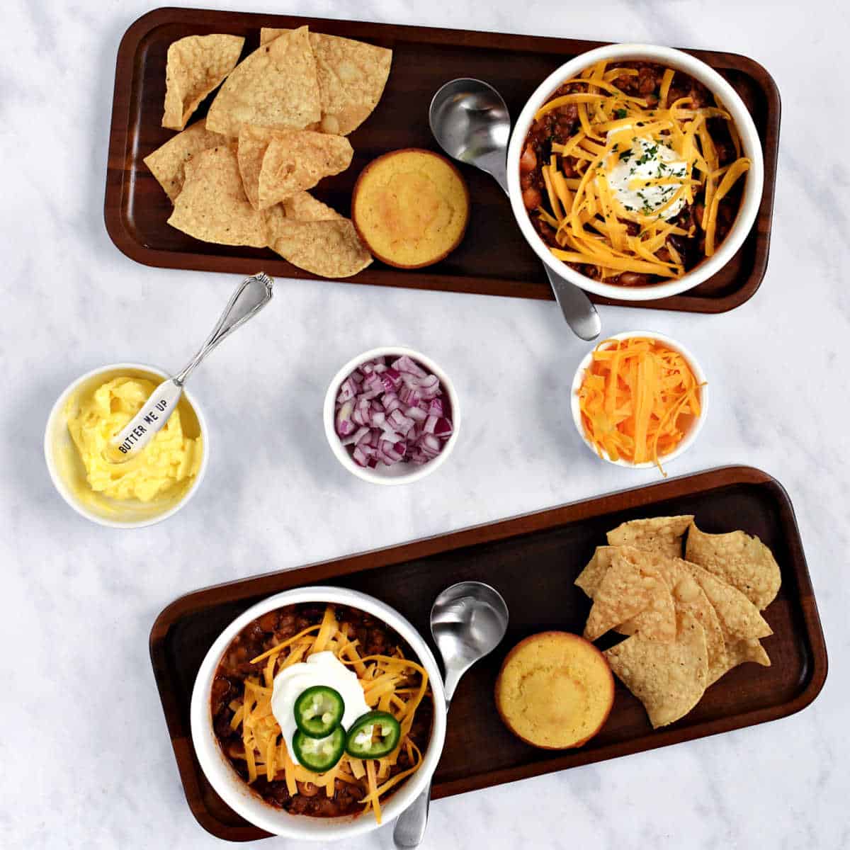 Bowls of chili, tortilla chips, corn muffins, and toppings on platters.