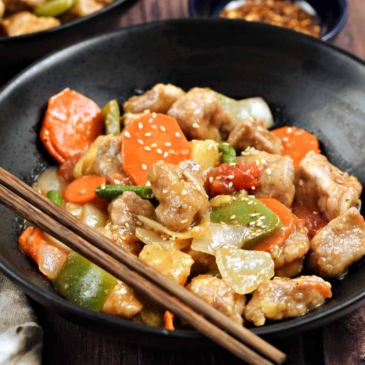 Sweet and sour pork with pineapple, carrots, onion, bell pepper, green onions in a bowl with chopsticks on the edge.