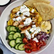 Sliced chicken, cucumber, cherry tomatoes, diced red onion, hummus, and yellow rice in a bowl and drizzled with yogurt sauce.