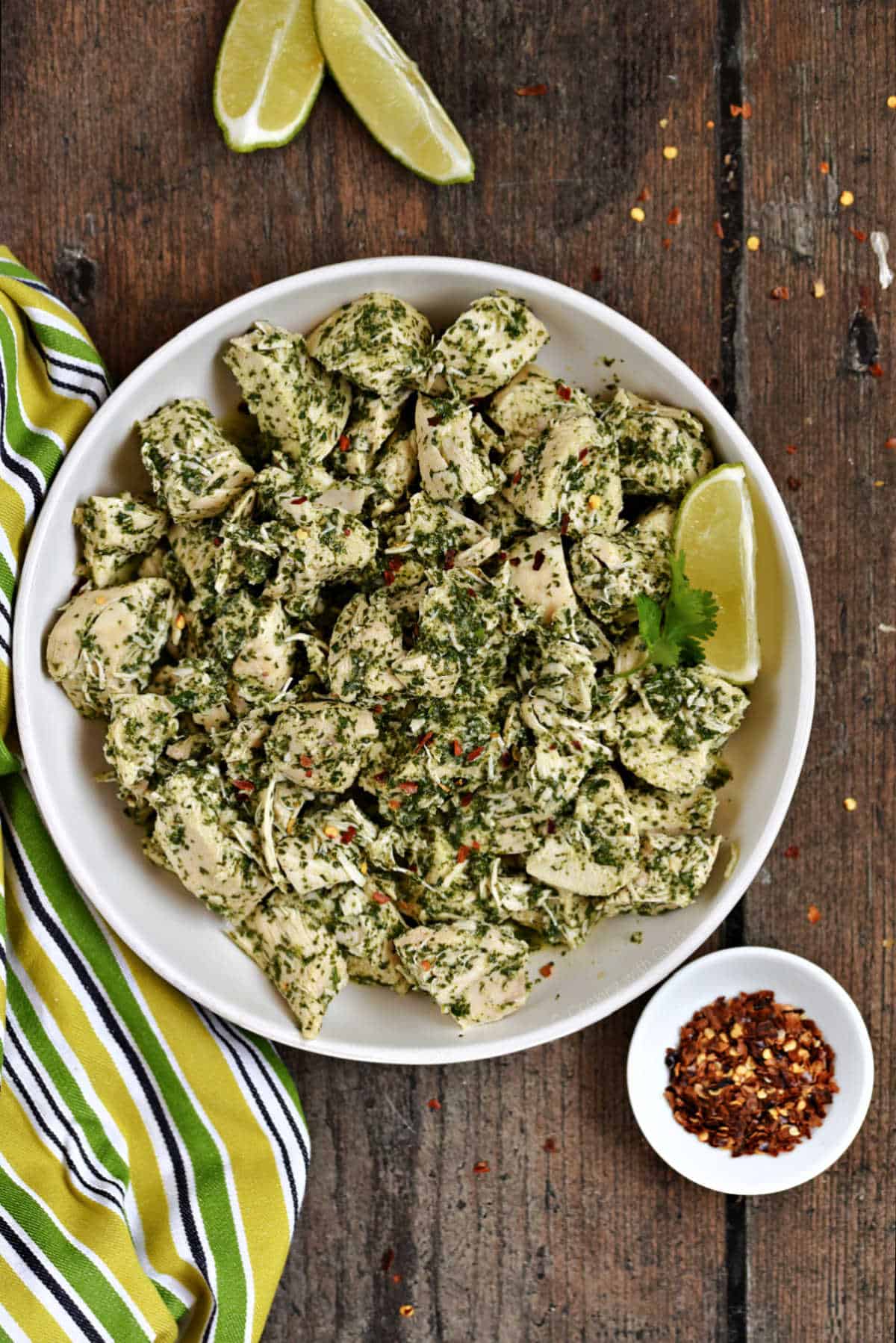 Chimichurri chicken in a serving bowl with red pepper flakes and lime wedges on the side.