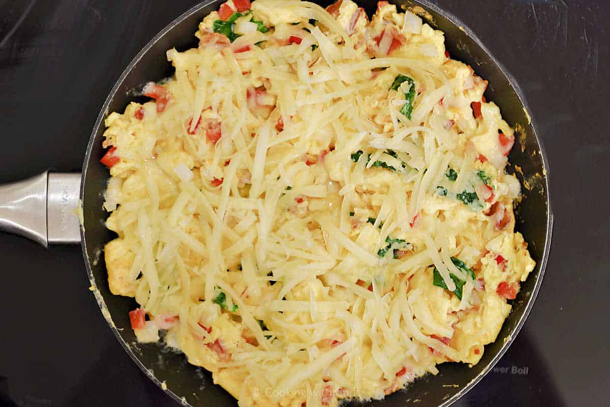 Shredded cheese over scrambled eggs, spinach, peppers, and onion in a skillet.