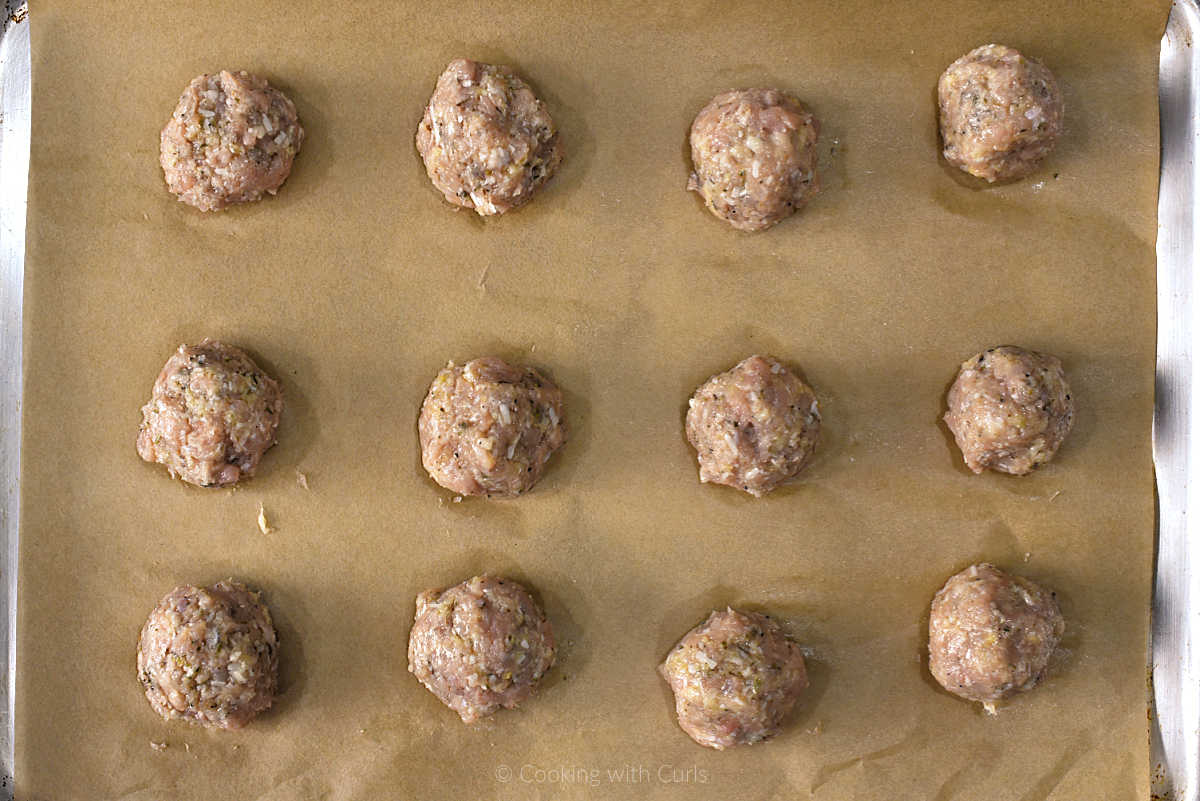 Twelve raw chicken meatballs on parchment lined baking sheet.