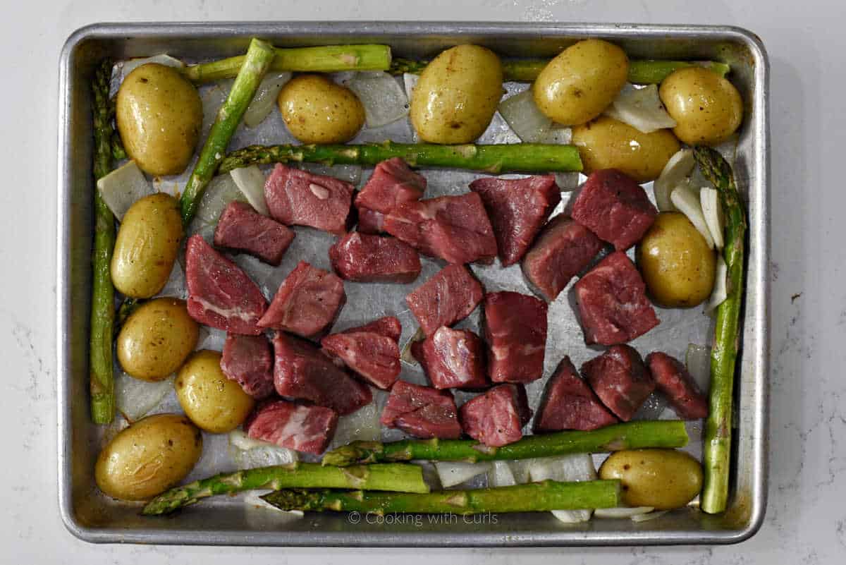 Raw steak cubes, potato halves, and asparagus spears on sheet pan.