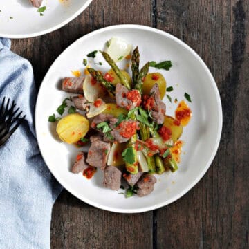 Steak bites, gold potato halves, onion, and asparagus spears topped with tomato butter and chopped cilantro on a plate.