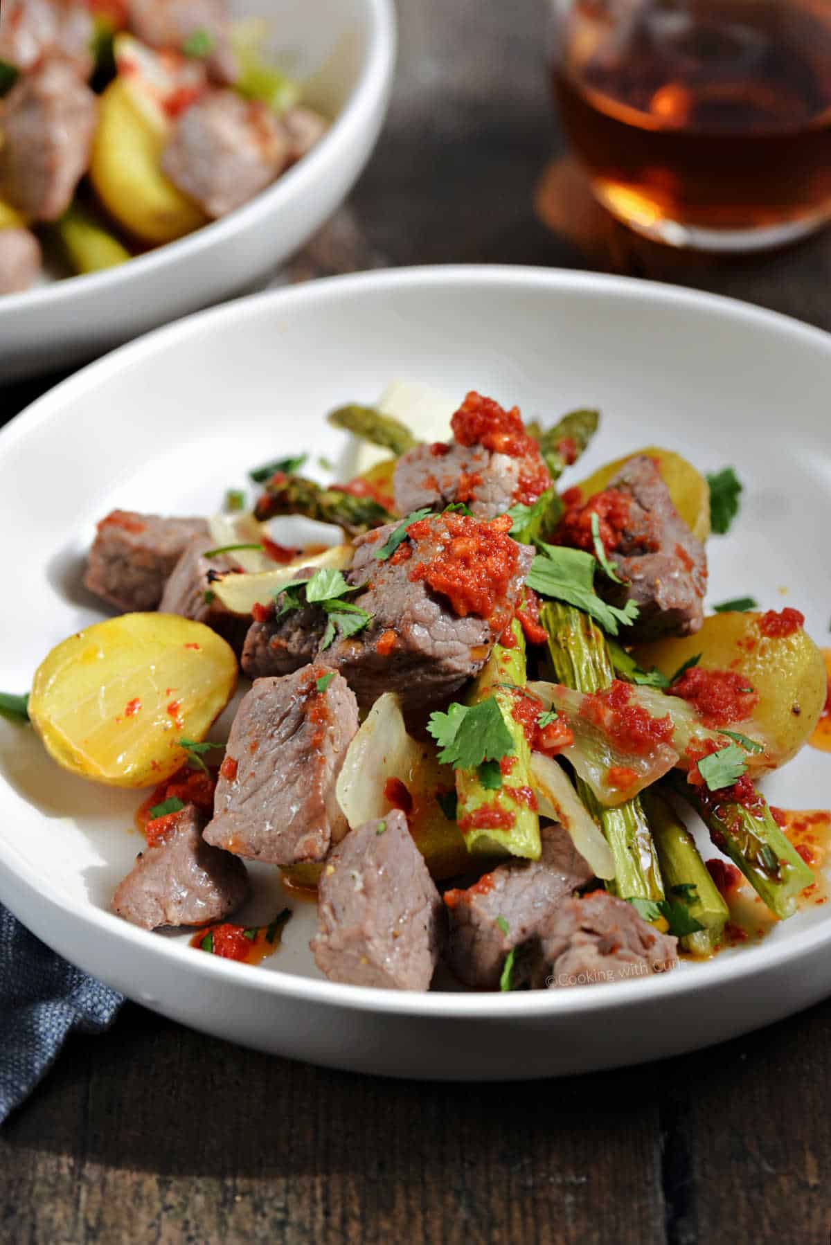 Steak bites, gold potato halves, onion, and asparagus spears topped with tomato butter and chopped cilantro on a plate.